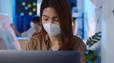 Mutlu Asyalı iş kadını, iş yerinde dizüstü bilgisayar kullanırken, virüs önleme amacıyla sosyal mesafeler için tıbbi maske takıyor. Coronavirüs 'ten sonra hayat ve iş.