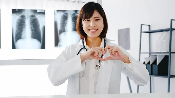 Confident young Asia female doctor in white medical uniform making a heart gesture with her fingers and smiling while video conference call with patient in hospital. Consulting and therapy concept.
