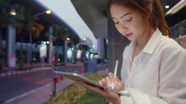 Moda bürosu kıyafetleri içinde başarılı genç Asyalı iş kadını dijital tablete yazmak için akıllı kalem teknolojisi kullanırken gece şehir çağında açık havada tek başına oturuyor. İş konsepti..