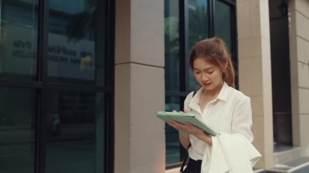 デジタルタブレットを使用したファッションオフィスの服の若いアジアのビジネスマンの成功と朝の都会の近代的な都市で屋外で一人歩きしながらテキストメッセージを入力します 外出先の概念上のビジネス — ストック動画