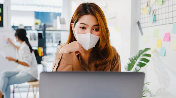 Asia businesswoman wear face mask for social distancing in new normal situation for virus prevention while using laptop presentation to colleagues about plan in video call while work in office.
