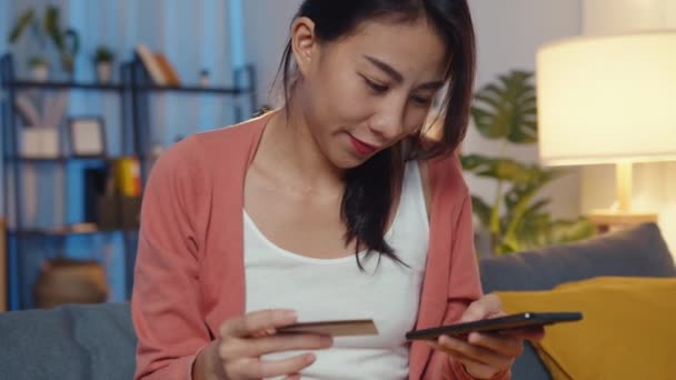 夜の時間美しい笑顔アジアの女性はリビングルームでソファの上にクレジットカードでオンラインショッピング製品を注文携帯電話を使用しています 自宅での滞在 自己隔離活動 居心地の良い隔離のための楽しい活動 — ストック動画