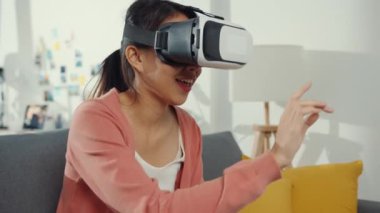 Sanal gerçeklik gözlüğü takan Asyalı kadın evdeki oturma odasında kanepede oturuyor. Evde kal, karantinada kal, gerçekliği yeniden hayal et, gelecek konseptinin VR teknolojisi.