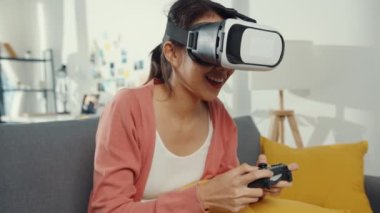 Asyalı kadın evdeki oturma odasındaki kanepede sanal gerçeklik gözlüğü takıp oyun oynuyor. Evde kalıp karantinaya alın, gerçekliği yeniden hayal edin, evde VR, gelecek konseptinin VR teknolojisi..