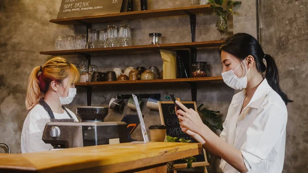Young Asia female wear face mask self service use mobile phone pay contactless at restaurant. Girl barista talking receive order from customer at cafe. Lifestyle new normal after corona virus concept.