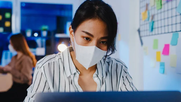 Happy Asia businesswoman wearing medical face mask for social distancing in new normal situation for virus prevention while using laptop back at work in office night. Life and work after coronavirus.