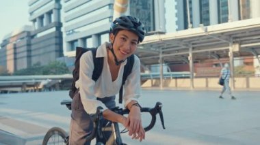 Asyalı iş kadını ofis standında işe gidiyor ve gülümsüyor sırt çantası takıyor bisiklet ile kameraya bakıyor ve bir şehrin etrafında bisiklet yapıyor. Bisiklet yolculuğu, bisikletle yolculuk, iş seyahati konsepti..