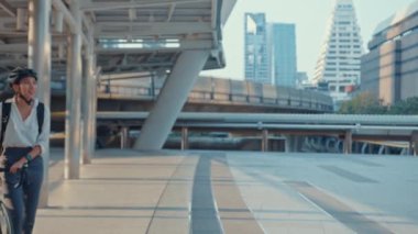 Asyalı iş kadını işe gider, işe gider, gülümser, sırt çantası takar, bisiklet sürer, şehir caddesinde bir bina inşa eder. Bisiklet yolculuğu, bisikletle yolculuk, iş seyahati konsepti..