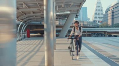 Asyalı iş kadını işe gider, işe gider, gülümser, sırt çantası takar, bisiklet sürer, şehir caddesinde bir bina inşa eder. Bisiklet yolculuğu, bisikletle yolculuk, iş seyahati konsepti..