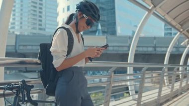 Sırt çantalı mutlu Asyalı iş kadını şehirde akıllı telefon kullanıyor bisiklet ile caddede duruyor işe gidiyor. Spor kızı iş için telefonunu kullanıyor. İşe gidip gelmek, şehirde iş seyahati yapmak..
