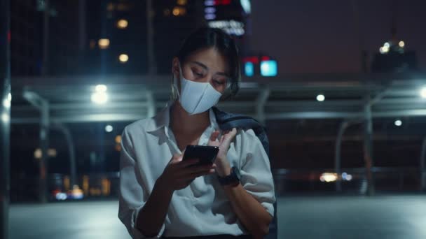 Junge asiatische Geschäftsfrau in modischer Kleidung trägt Gesichtsmaske und tippt per Smartphone SMS, während sie nachts im Freien in der Großstadt steht. Soziale Distanzierung zur Verhinderung der Verbreitung des COVID-19-Konzepts.