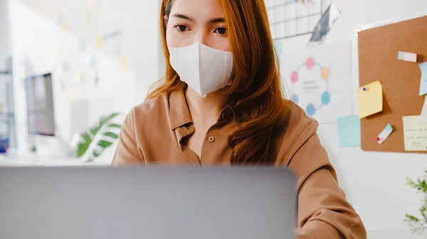 Asia businesswoman wear face mask for social distancing in new normal situation for virus prevention while using laptop presentation to colleagues about plan in video call while work in office.