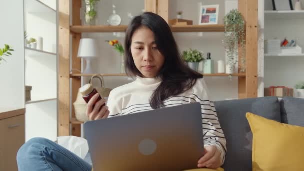 生病的年轻亚洲女人拿着药坐在沙发上 带着笔记本电脑在家里咨询医生 女孩下了医生的命令后才吃药 在家里被隔离 社会上远离了验尸官的概念 — 图库视频影像