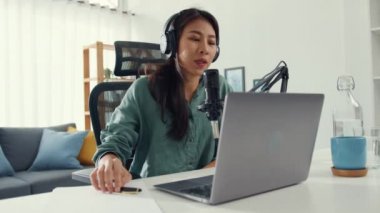 Mutlu Asyalı kız laptopuna kulaklıkla bir podcast kaydediyor ve odasında seyirciyle mikrofon konuşması yapıyor. Kadın podcaster kendi stüdyosundan ses yayını yapıyor. Ev konseptinde kalın..