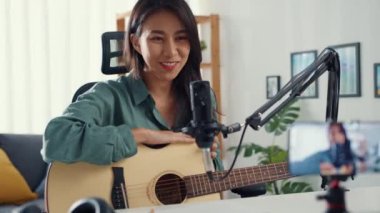 Genç Asya 'lı kız nüfuzlu biri gitar çalıyor. Evde dinleyiciler için akıllı telefonlu mikrofon kaydı kullanıyor. Kadın podcaster kendi stüdyosundan ses yayını yapıyor. Ev konseptinde kalın..