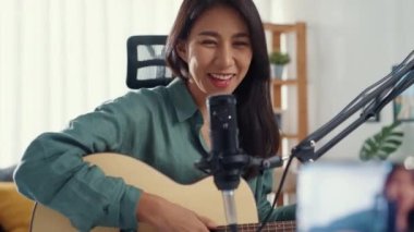 Genç Asya 'lı kız nüfuzlu biri gitar çalıyor. Evde dinleyiciler için akıllı telefonlu mikrofon kaydı kullanıyor. Kadın podcaster kendi stüdyosundan ses yayını yapıyor. Ev konseptinde kalın..
