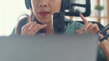 Mutlu Asyalı kız laptopuna kulaklıkla bir podcast kaydediyor ve odasında seyirciyle mikrofon konuşması yapıyor. Kadın podcaster kendi stüdyosundan ses yayını yapıyor. Ev konseptinde kalın..