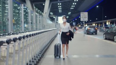 Asyalı iş kızı varış yerine giderken maske takıyor ve iç havaalanındaki garaj kapısının önünde kalkıyor. İş seyahati covid salgını, iş seyahati sosyal uzaklık kavramı.