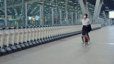 Asyalı iş kızı varış yerine giderken maske takıyor ve iç havaalanındaki garaj kapısının önünde kalkıyor. İş seyahati covid salgını, iş seyahati sosyal uzaklık kavramı.