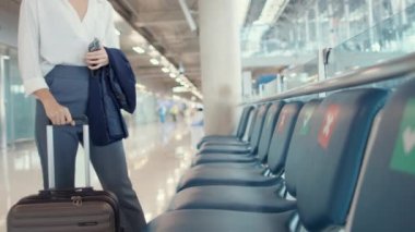 Asyalı iş kızı bavuluyla bankta oturup havaalanında uçuş partneri arıyor. İş seyahati covid salgını, iş seyahati sosyal mesafesi, iş seyahati konsepti.