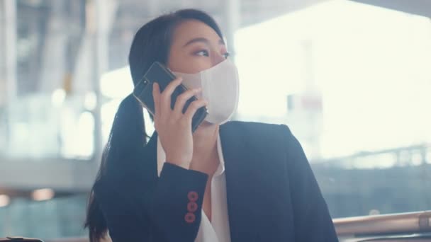 聪明的亚洲女商务人士穿着西服 坐在长椅上提着手提箱 与合作伙伴通电话 等待机场的航班 大流行病中的商务旅行通勤者 商务旅行概念 — 图库视频影像