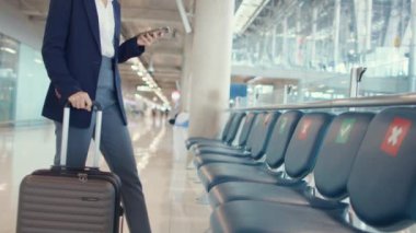 Asyalı iş kadını seyahat eden bayan bavulla oturan takım elbiseli ve bankta akıllı telefon sohbet mesajı kullanarak havaalanında uçuşu bekliyor. İş seyahati gezisi salgını, iş seyahati konsepti.