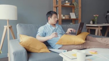 Serbest Asyalı adam evdeki oturma odasında dizüstü bilgisayar kullanarak günlük kıyafet giyiyor. Evden çalışmak, uzaktan çalışmak, uzaktan eğitim, sosyal uzaklık, korona virüsünün önlenmesi için karantina.