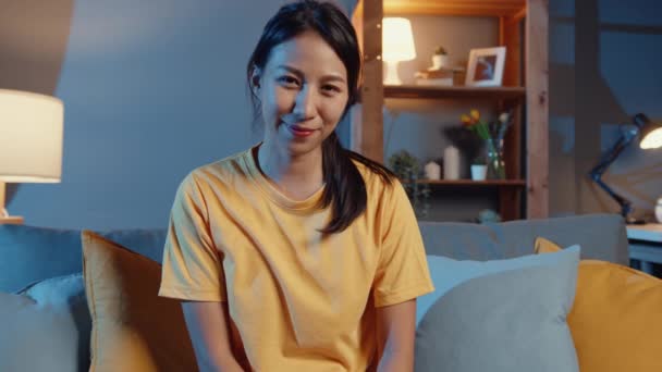 快乐的年轻亚洲自由职业女性 看着相机微笑着 愉快地在网上视频里放松一下 晚上呆在客厅里 待在家里隔离 在家工作 与他人保持距离 — 图库视频影像
