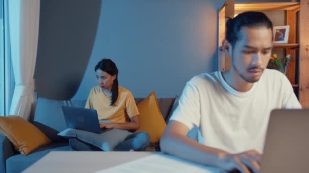 フリーランスのアジアのカップルの男性と女性のカジュアルな仕事に真剣に集中してノートパソコンの夜には 夫のテーブルの妻の前に座ってソファの上に家の後ろに座っている 家のコンセプトからの仕事 — ストック動画