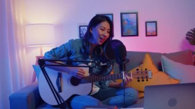 Mutlu Asyalı kız blogcu gitar çalar ve mikrofon çalar müzik çalar ses mikserini modern oturma odasındaki ev stüdyosunda gece yarısı dizüstü bilgisayarında kullanır. Müzik içeriği oluşturucu, Öğretici, Yayın konsepti.