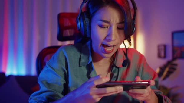 幸せなアジアの女の子のゲーマーは 自宅のリビングルームでスマートフォンのカラフルなネオンでオンラインビデオゲームをプレイヘッドフォン競争を着用してください スポーツストリーミングゲーム ホーム隔離活動の概念 — ストック動画