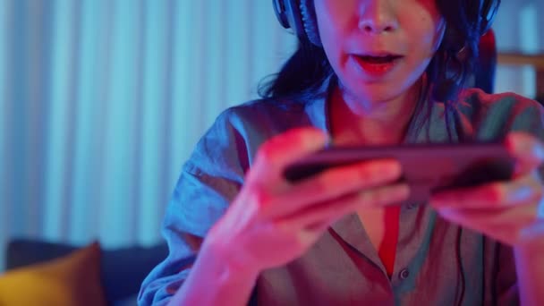 幸せなアジアの女の子のゲーマーは 自宅のリビングルームでスマートフォンのカラフルなネオンでオンラインビデオゲームをプレイヘッドフォン競争を着用してください スポーツストリーミングゲーム ホーム隔離活動の概念 — ストック動画
