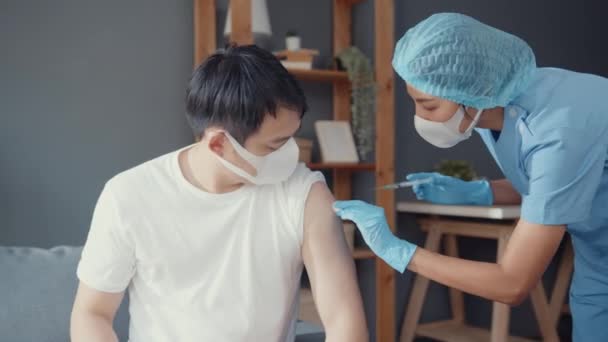男性患者に接種したCovid 19またはインフルエンザウイルスワクチンを投与した若いアジアの女性看護師は 自宅のリビングルームでソファの上に座っているウイルス病から顔マスク保護を着用してください 予防接種の概念 — ストック動画