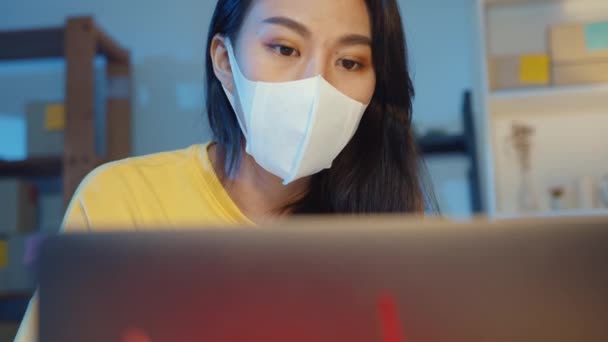 Fiatal ázsiai üzletasszony visel arc maszk ellenőrzés megrendelés és válasz ügyfél postaláda chat laptop otthoni irodában éjszaka. Kisvállalkozás tulajdonos, online piaci szállítás, életmód szabadúszó koncepció.