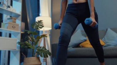 Spor kıyafetli genç Asya 'lı bayan, kollarını gerdirme egzersizinde halterlerle çömelmiş, geceleri oturma odasında çalışıyor. Spor ve eğlence aktivitesi, korona virüsü konsepti için karantina.
