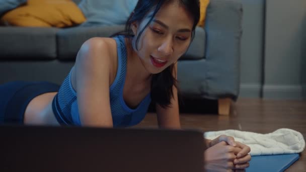 スポーツウェアの練習をしている若いアジアの女性は 自宅の夜にヨガビデオチュートリアルを見るためにラップトップを使用して動作します パーソナルトレーナーとの遠距離トレーニング 社会的距離 オンライン教育の概念 — ストック動画