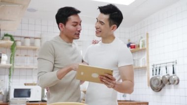 Asyalı eşcinsel çift tablet kullanıyor ve kahvaltı hazırlıyor. Evde mutfaktaki masada sebzeli sandviç. Genç LGBT erkekleri mutlu konuşuyorlar. Birlikte dinleniyorlar. Sabahları evde romantik vakit geçiriyorlar..