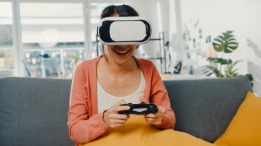 Asyalı kadın, evdeki oturma odasındaki kanepede sanal gerçeklik gözlüğü takıp joystick oynuyor. Evde kalıp karantinaya alın, gerçekliği yeniden hayal edin, evde VR, gelecek konseptinin VR teknolojisi..