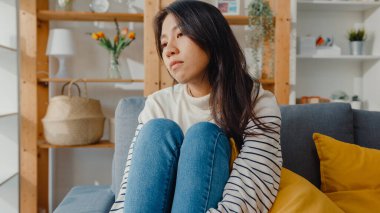 Düşünceli Asyalı kadın oturma odasında dizlerini kucaklayarak oturur kendini yalnız hisseder, üzgün genç kız evde yalnız vakit geçirir, sosyal mesafe, Coronavirüs karantinası..