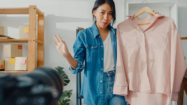 Young Asia lady fashion designer using mobile phone receiving purchase order and show clothes recording video live streaming online with camera. Small business owner, online market delivery concept.