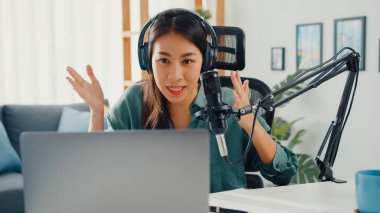 Mutlu Asyalı kız laptopuna kulaklıkla bir podcast kaydediyor ve odasında seyirciyle mikrofon konuşması yapıyor. Kadın podcaster kendi stüdyosundan ses yayını yapıyor. Ev konseptinde kalın..