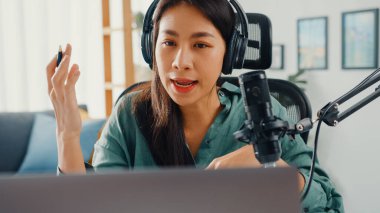 Mutlu Asyalı kız laptopuna kulaklıkla bir podcast kaydediyor ve odasında seyirciyle mikrofon konuşması yapıyor. Kadın podcaster kendi stüdyosundan ses yayını yapıyor. Ev konseptinde kalın..