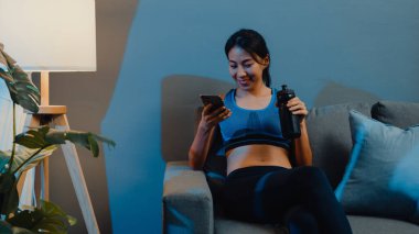 Genç Asyalı bayan cep telefonu kullanıyor ve evdeki oturma odasındaki koltukta saf su içiyor. Spor ve eğlence aktivitesi, sosyal uzaklık, korona virüsü önleme konsepti için karantina.
