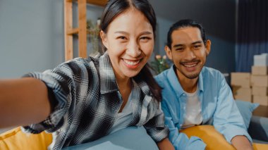 Mutlu genç Asyalı çift kanepede oturur ve evdeki aile ve arkadaşlarıyla kamera görüntülerini izlerler. Evde karantina, sosyal mesafeler, genç evlilik kavramı..