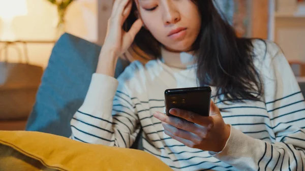 Thoughtful Asia lady holding phone feeling sad waiting for call sit at sofa in living room at house night feel lonely, Sad depressed teenager spend time alone, Social distance, Coronavirus quarantine.