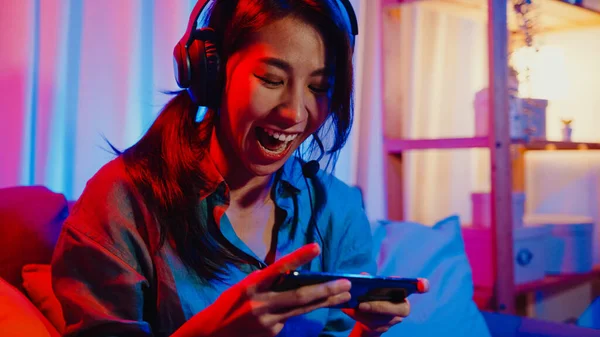 幸せなアジアの女の子のゲーマーは 自宅でカラフルなネオンのリビングルームでソファの上に座っている友人とスマートフォンの興奮話とオンラインヘッドフォン競争ビデオゲームを着用し ホーム隔離活動のコンセプト — ストック写真