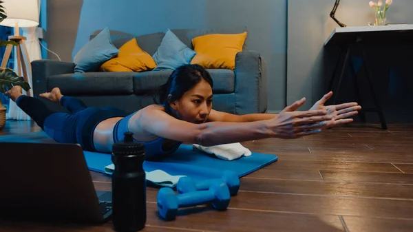 Young Asia lady in sportswear exercises doing work out and using laptop to watch yoga video tutorial at home night. Distant training with personal trainer, social distance, online education concept.