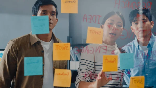 Asia businesspeople stand behind transparent glass wall listen manager pointing progress work and brainstorm meeting and worker post sticky note on wall at office. Business inspiration, Share ideas.