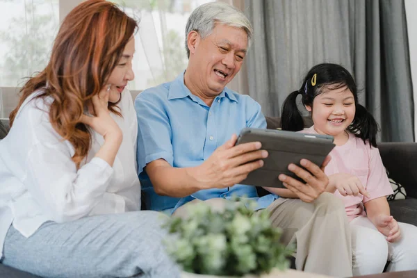 Asian grandparents and granddaughter using tablet at home. Senior Chinese, grandpa and grandma happy spend family time relax with young girl checking social media, lying on sofa in living room concept