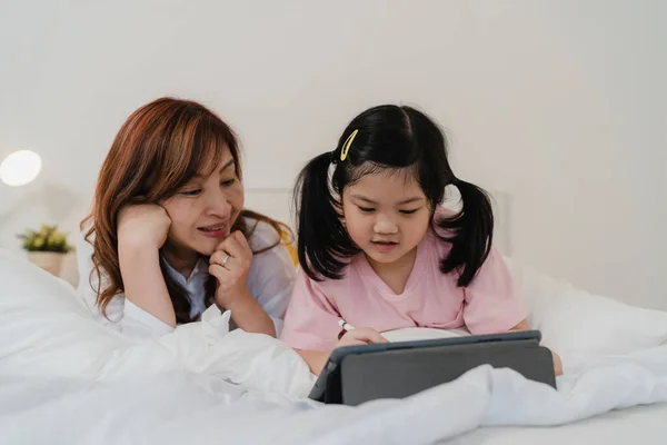 Asian grandmother using tablet read fairy tales to granddaughter at home. Senior Chinese, grandma happy relax with young girl before bedtime lying on bed in bedroom at home at night concept.